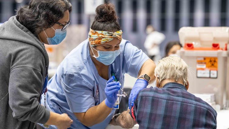 Nurse in blue scrubs giving a vaccine to a senior citizen as someone else looks on