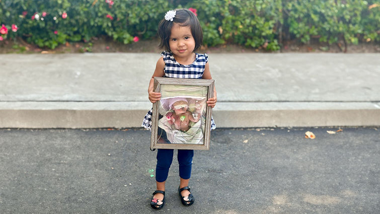 Little girl wearing black and white dress with leggings and flats holds a photo of herself when she was in the NICU