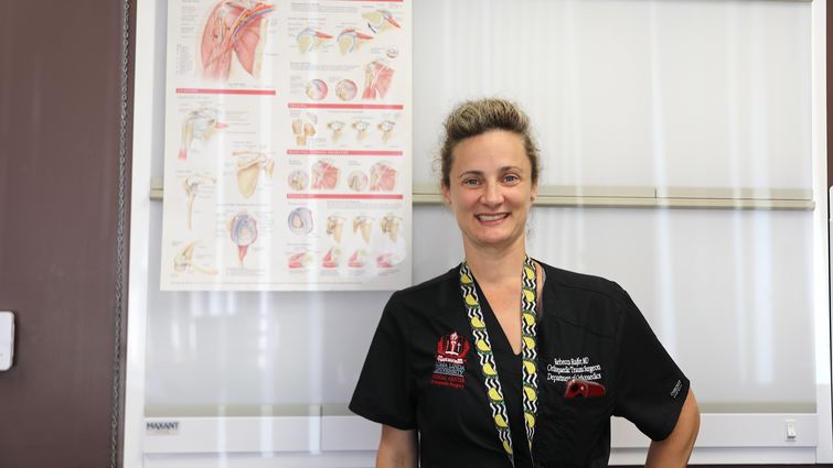 Dr. Rajfer stands next to a poster of the shoulders anatomy as she smiles at the camera