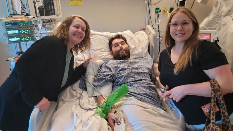 Two woman lean over a man in a hospital bed while all three smile at the camera