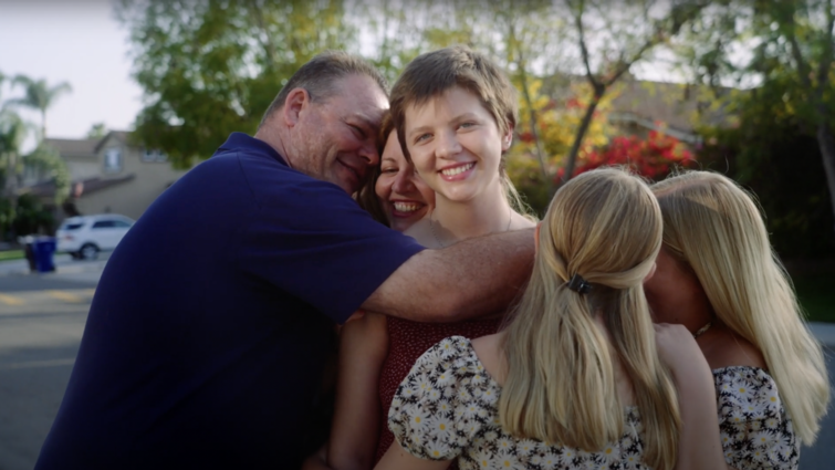 family surrounds teenage daughter, all in a group hug outside smiling broadly
