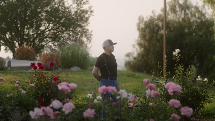 Young hispanic stands in garden with arms relaxed behind his back. 