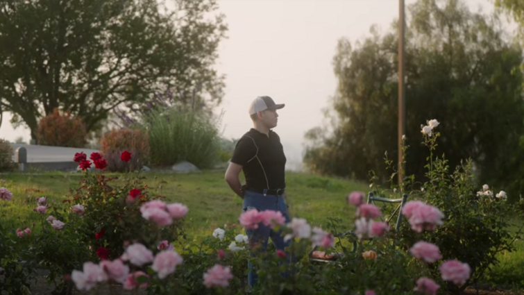 Young hispanic stands in garden with arms relaxed behind his back. 