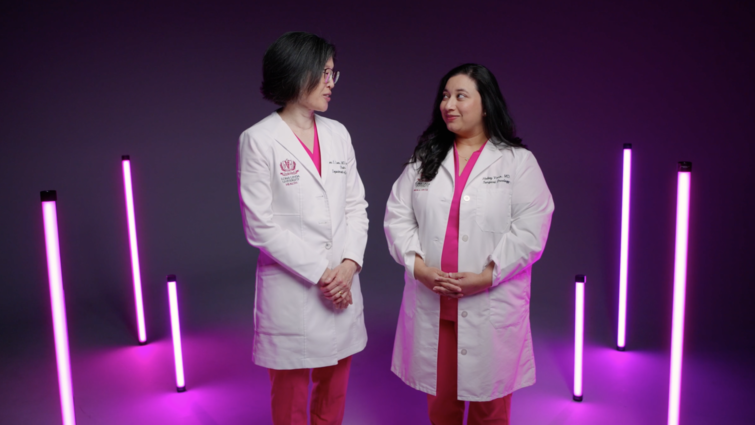 Two women stand shoulder to shoulder with pink light fixtures standing behind them. Both women are wearing pink scrubs with white doctors coats. Woman on left is Asian and woman on right is middle-eastern. 