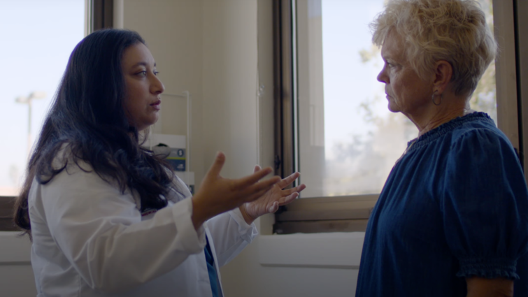 Doctor with brown hair in white coat has a conversation with a patient wearing blue top with short blonde hair. 