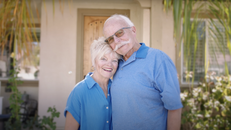 Elderly caucasian couple stands smiling side by side both wearing light blue. 