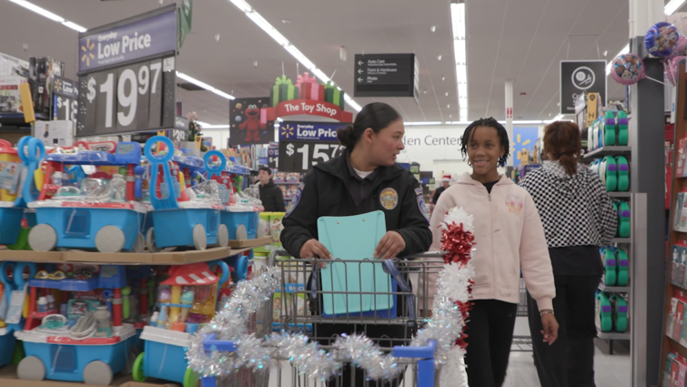 A female officer walks alongside a pediatric RICA patient in Walmart with a Christmas decorated shopping cart filled with toys.