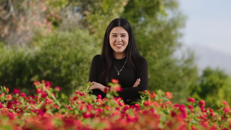 Young woman with dark hair and a black shirt stands smiling with her arms crossed in front. Red flowered bushes are in the foreground. 