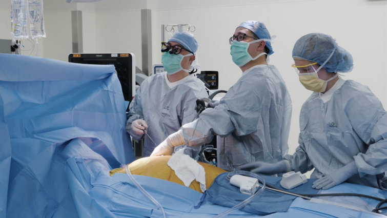 Three people in blue scrubs stand over operating table looking to the viewers left.