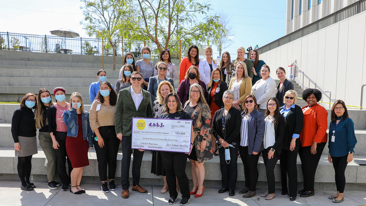 group of child life specialists and hospital administrators stand with giant check in outdoor amphitheater smiling at camera