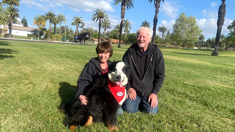 Husand and wife sit in a field with their therapy dog named Emma