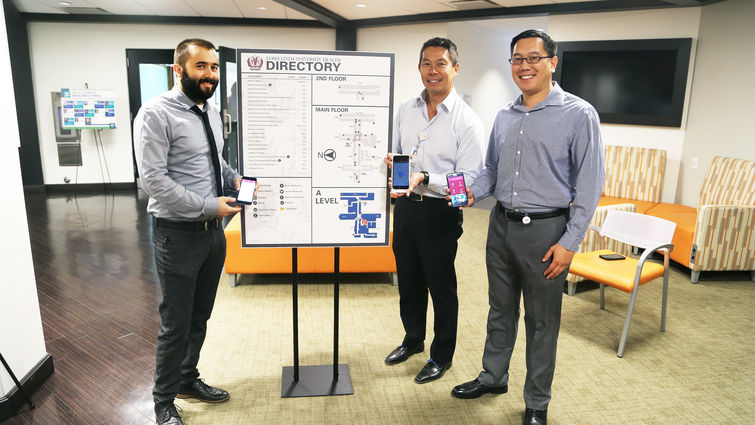 Standing by a traditional directory and map of Nichol Hall from left to right, Marcus Chapman, Kent Chow and Nicholas Yu display the new mobile wayfinding app that is now being used by faculty, students and staff to help navigate the historic building.