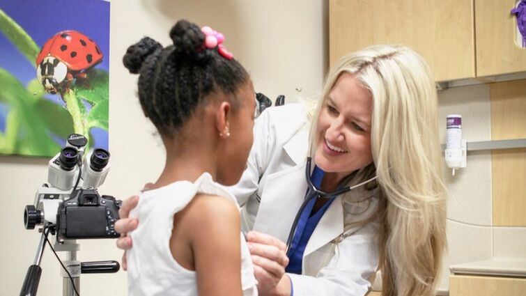 A healthcare provider wearing a white coat uses a stethoscope to examine a child seated on an exam table in a pediatric clinic. Medical equipment and cabinets are visible in the background.