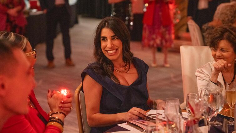 Guests sit at an elegantly set table during a gala event, surrounded by warm lighting, glassware, and other attendees in the background.