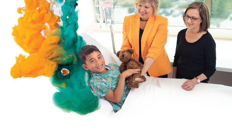 two women stand next to patient's bed giving him teddybear