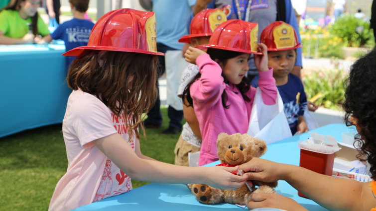 Children wearing red toy firefighter helmets gather around an outdoor activity table where an adult helps one child use a syringe on a stuffed teddy bear as part of a hands-on demonstration