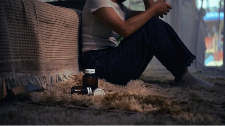 teen sits on floor by bed with bottle of pills closeby