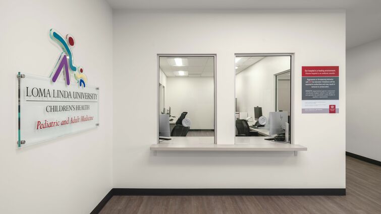 Reception area at Loma Linda University Children’s Health Pediatric and Adult Medicine clinic with two service windows, a counter, office workstations visible behind glass, and clinic signage on a bright, modern interior wall.