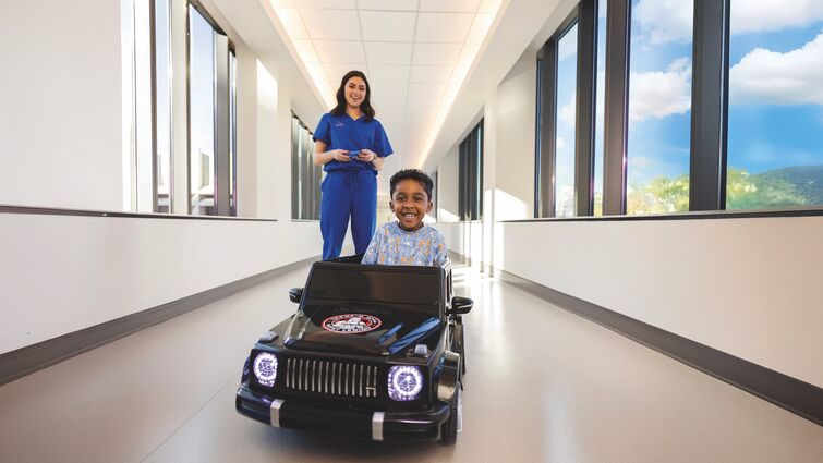 woman in royal blue scrubs standing behind children's hospital patient wearing hospital gownin a toy car