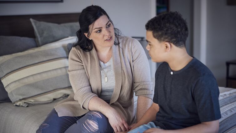 A Caucasian mother talking with mixed-race son while both sit on a bed