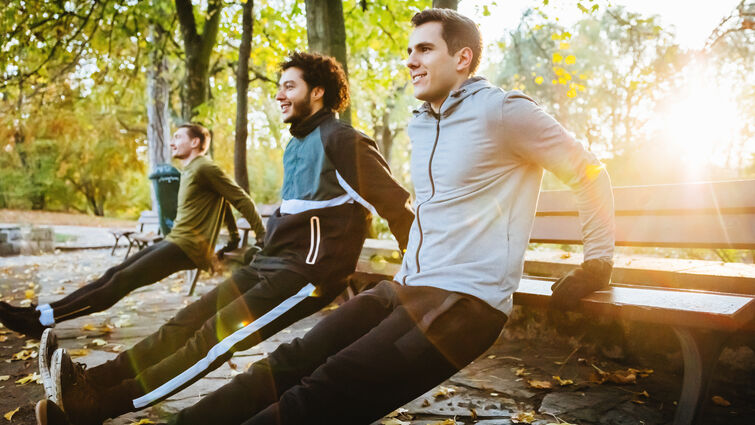 young men are leaning on a bench and stretching in a wooded forest 