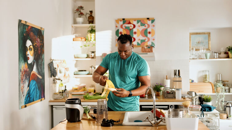 An African-American male in a bright kitchen prepares a banana at a wooden countertop surrounded by cooking tools, fresh produce, and a blender, with colorful artwork and neatly arranged shelves in the background.
