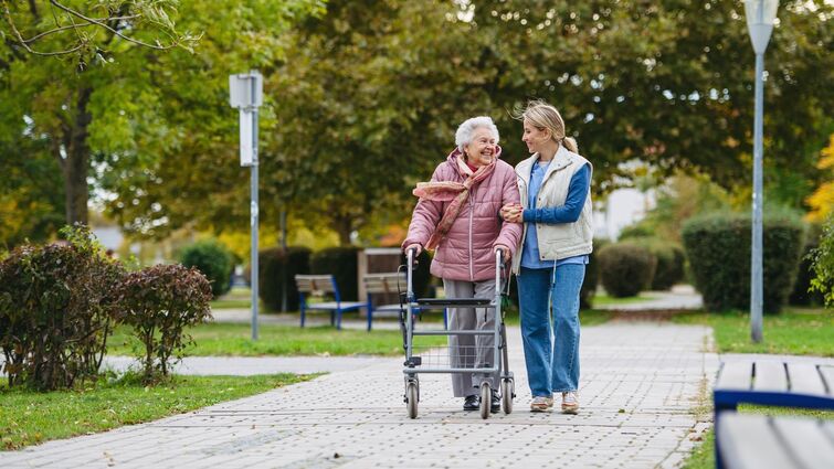 Senior woman and her home caregiver spending a chilly, windy day outdoors in city park. Autumn walk for elderly patient with walker