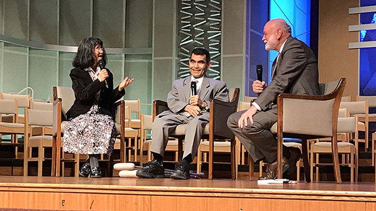 Two men and a woman sit in chairs on stage holding microphones