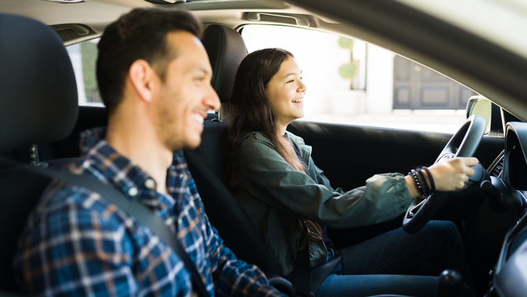 smiling father in passenger seat while teenage daughter sits in drivers seat with two hands on wheel, smiling