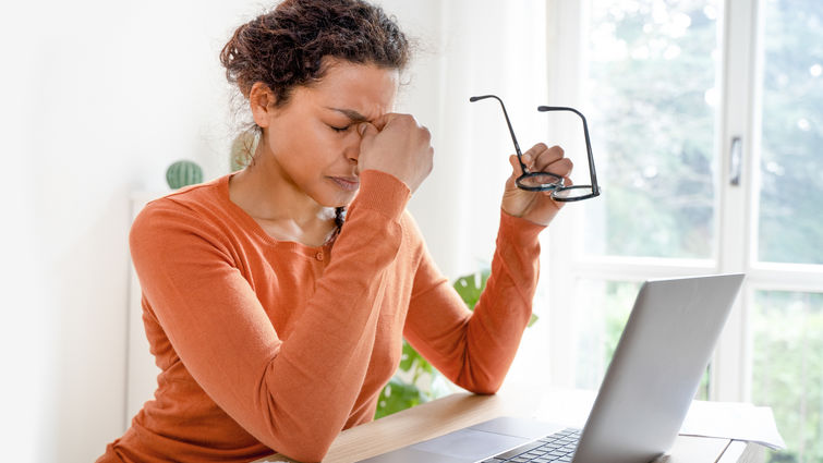 Woman sitting, pinching top of her nose holding eye glasses in front of a laptop