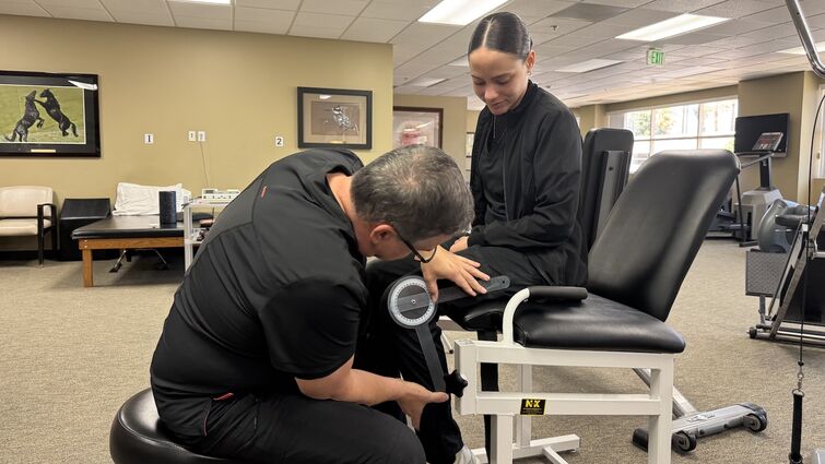 PT is essential in rehabbing old and new injuries, as well as aiding surgery recovery. A therapist in black scrubs leans over a female patients knee as she sits in a chair