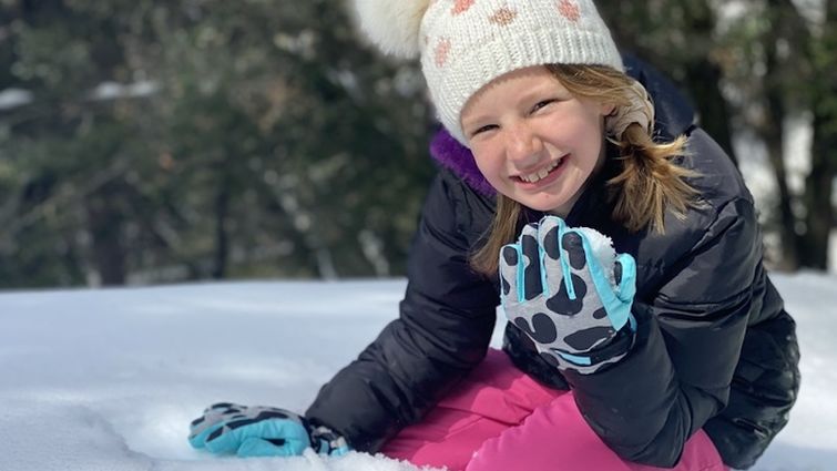 Young girl in beanie sitting on snow making snowballs