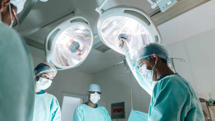 Four health workers wearing surgery clothes and masks, standing face to face in operation room with lamps on top.