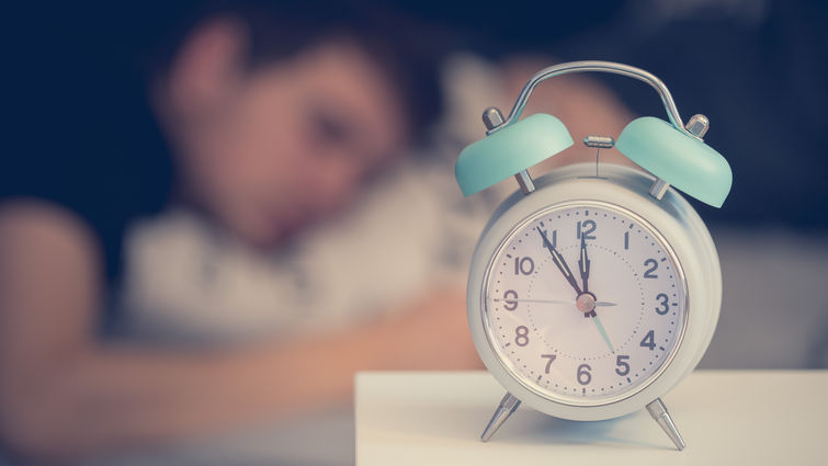 Alarm clock on nightstand in foreground, person sleeping in the background