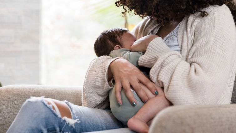 Sitting on a chair in her living room, the unrecognizable mom holds her baby snugly while breastfeeding him
