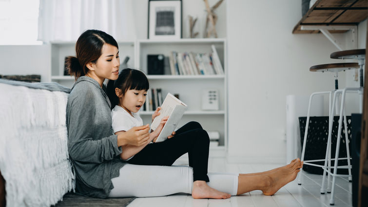 Young Asian mother sitting on the floor in the bedroom reading book to little daughter, enjoying family bonding time together at home