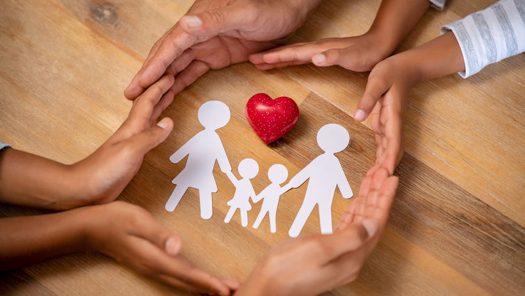 Close up hands of father, mother and daughters protecting family paper cutout with red heart. Hands of mixed race family with paper cutting and red heart shaped symbol stone on wooden background.