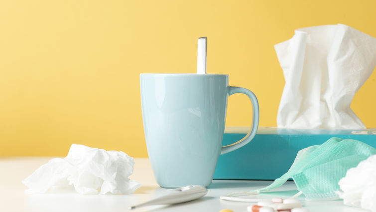Tissue box, tissues, thermometer, protective face mask, capsules and a hot drink on white table in front of yellow wall.