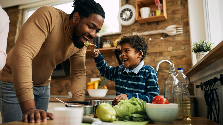 Happy African American boy having fun while feeding his father in the kitchen.