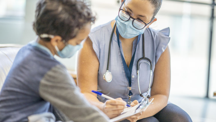 A Hispanic female doctor is taking down notes while speaking to a young 8 year old male patient. Both doctor and patient are wearing masks during the coronavirus pandemic.