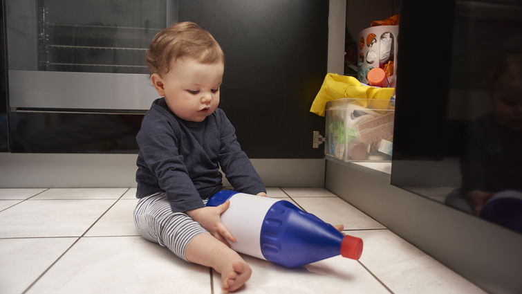 child sitting on the floor holding household chemical bottle from under the sink cabinetry