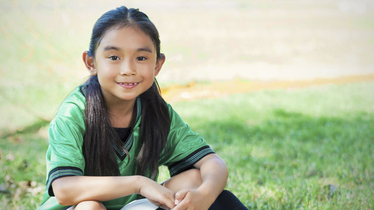 elementary age Hispanic female soccer player sits on the ground after her game. She is smiling big and holding a soccer ball. She has long brown hair and brown eyes. The playing field and soccer goal are in the background.