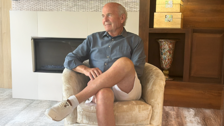White man in a blue button up shirt and white shorts sits in a yellow chair in front of a fireplace