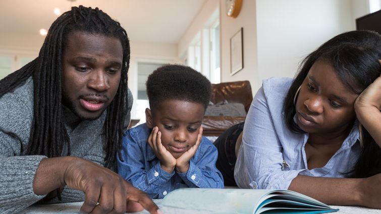 A preschool-age African American boy squeezes between his parents for reading time. They are sitting and sprawled out in the living room. He is resting his chin in both hands.