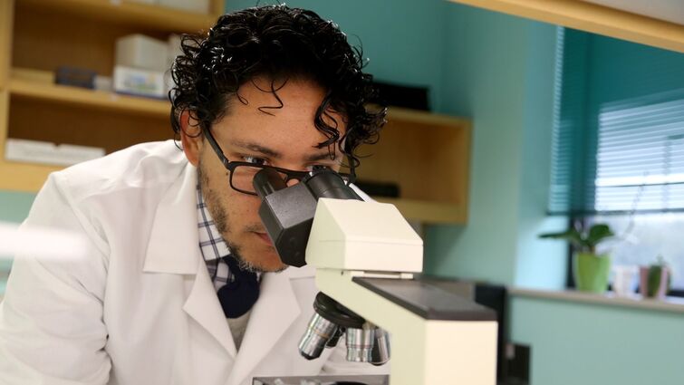 A doctor in a white lab coat and black hair looks through a microscope