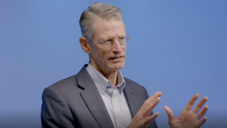a man in front of a blue background is talking with his hands up 