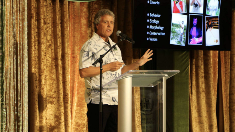 Bill Hayes standin on stage behind a podium with a presentation screen to the side.