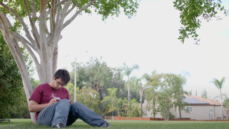 Tyler Rojanaroj sits at the base of a tree writing music.