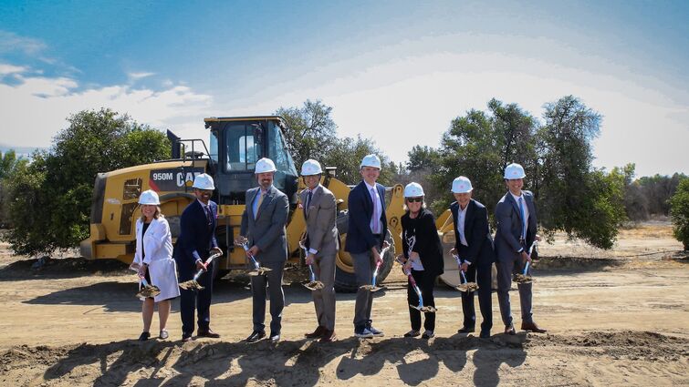 eight hospital leadership representatives dig dirt during the groundbreaking ceremony in front of a dump truck with trees in the background.
