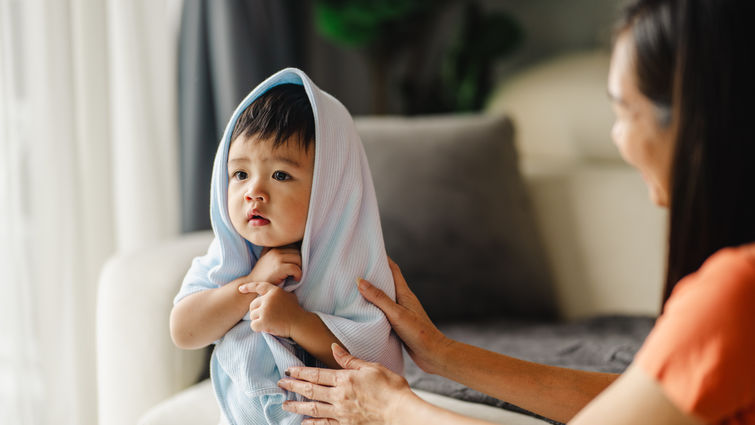 Asian boy with a towel on his head is sitting in the living room. He has big, dark black eyes and is relaxing on the sofa after his bath.